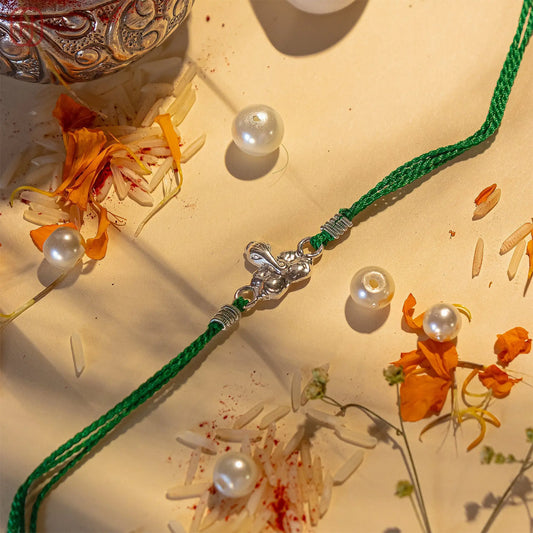 925 silver Ganesh Ji Rakhi with green thread and pearls surrounded by flowers and rice grains