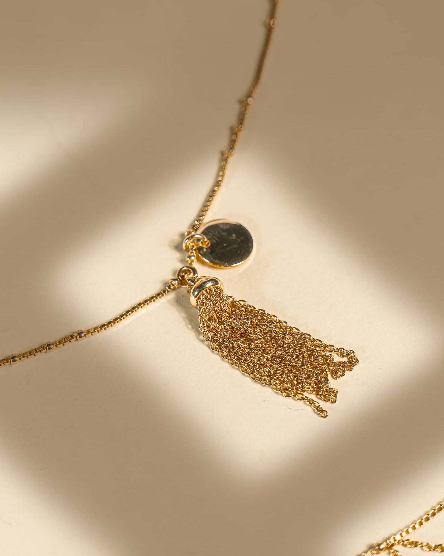 Pair of 18K gold plated sterling silver anklets featuring a dangling chain tassel and disc charm on a beige background.