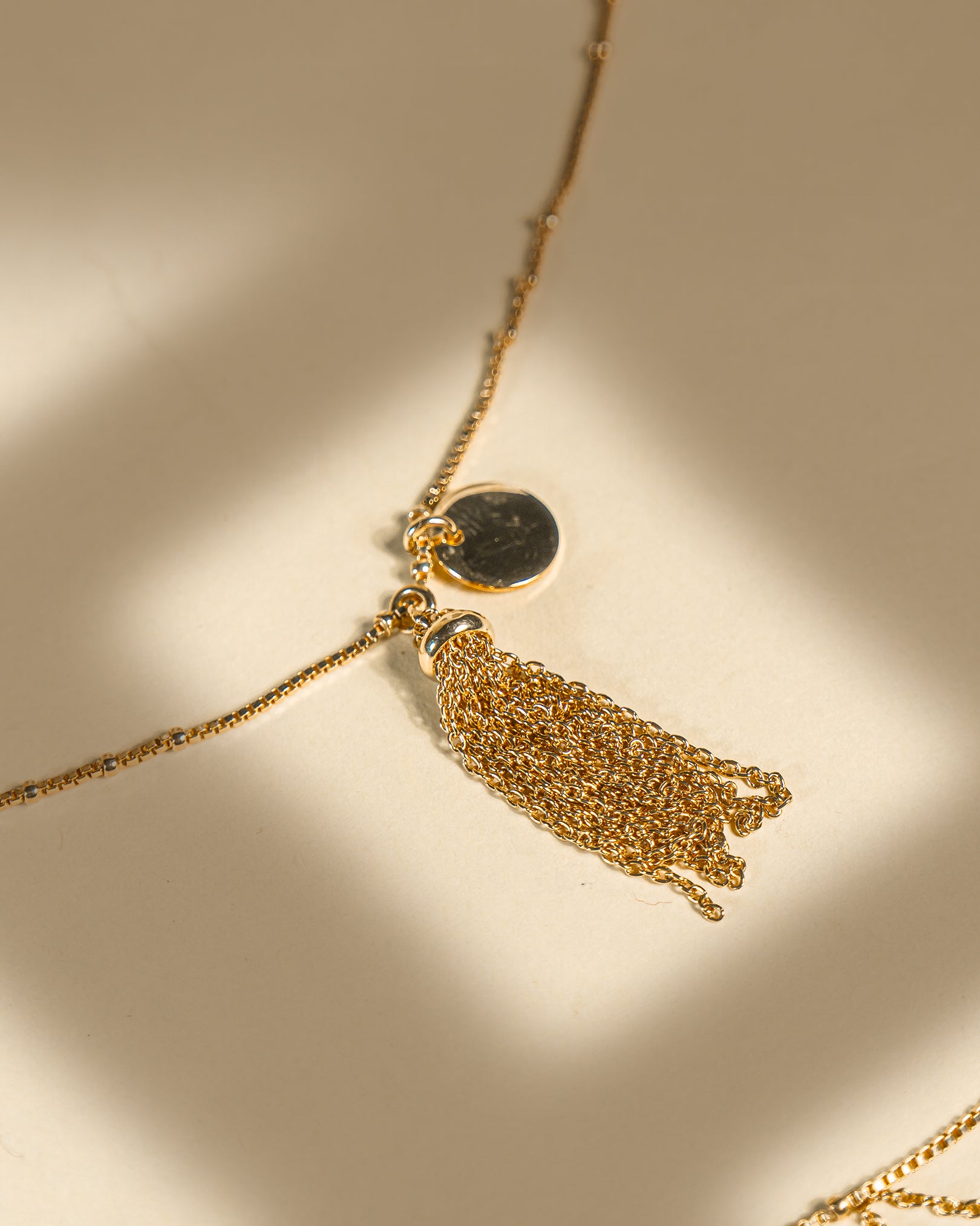 Pair of 18K gold plated sterling silver anklets featuring a dangling chain tassel and disc charm on a beige background.