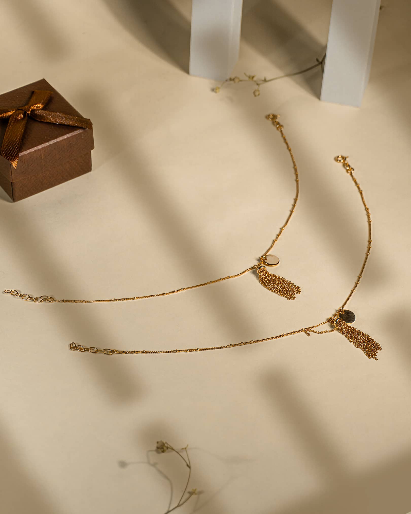 Pair of 18K gold plated sterling silver anklets featuring a dangling chain tassel and disc charm on a beige background.