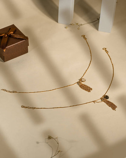 Pair of 18K gold plated sterling silver anklets featuring a dangling chain tassel and disc charm on a beige background.