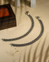 Pair of traditional oxidized silver anklets featuring repeating paisley mango motifs and small silver beads on a beige background.
