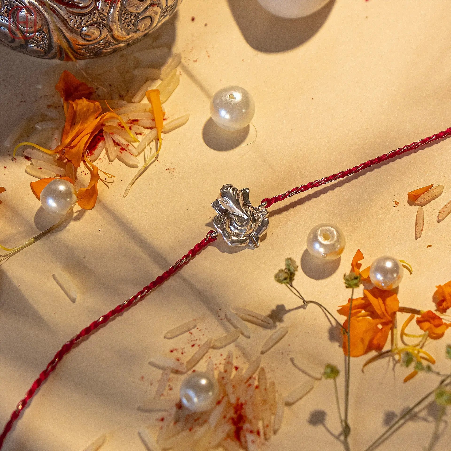 Ganesh silver rakhi with red thread surrounded by white pearls and marigold petals on a beige surface