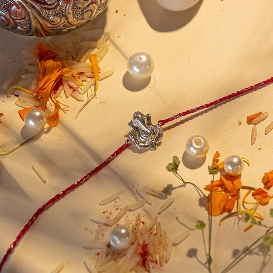 Ganesh silver rakhi with red thread surrounded by white pearls and marigold petals on a beige surface