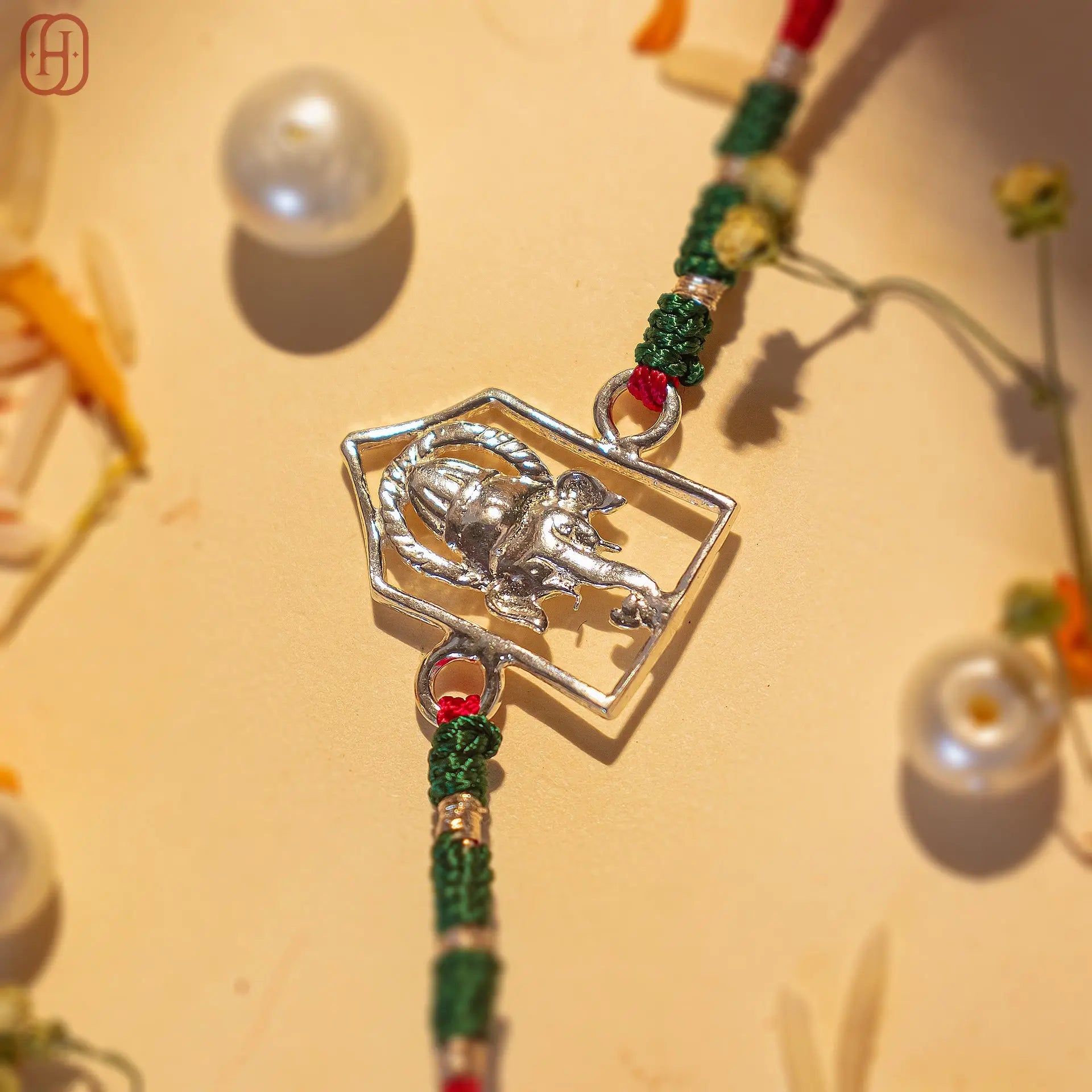 Silver Ganesh Ji face rakhi online with green and red thread by Hardik Jewels on beige background