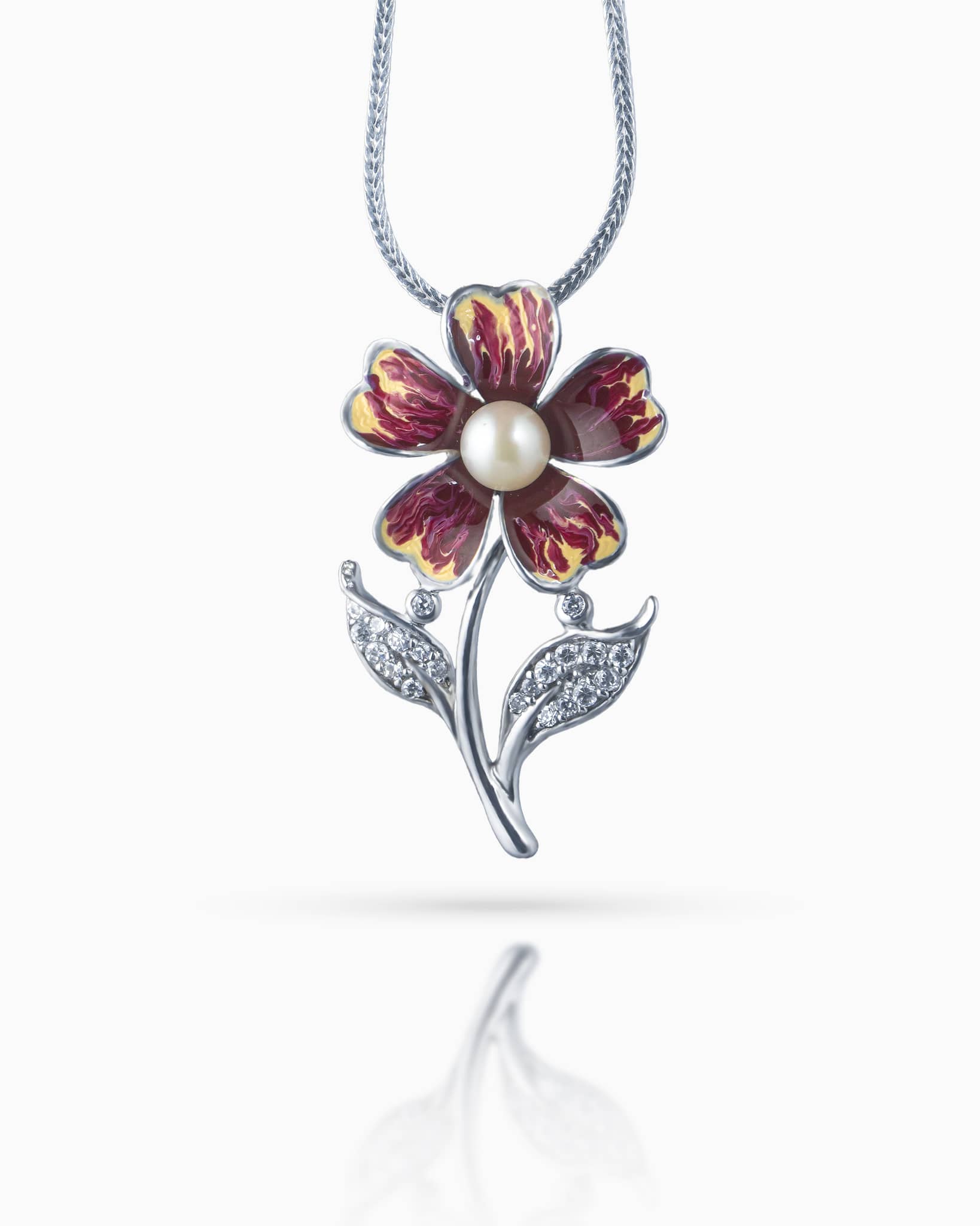 Close up of an elegant silver floral pendant showing detailed maroon and yellow enamel work, a white pearl, and cz paved leaf accents resting on a white quilted surface.