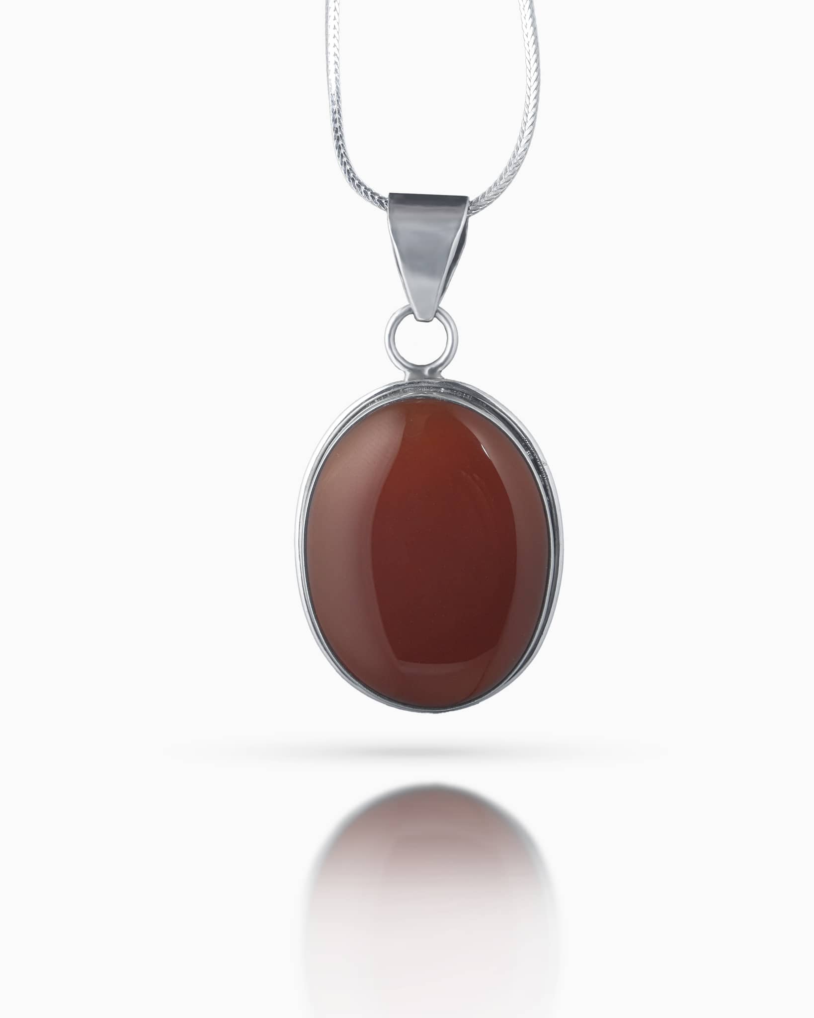 Close up of an elegant silver pendant showing a smooth, large oval reddish brown stone set in a simple silver bezel resting on a white quilted surface.