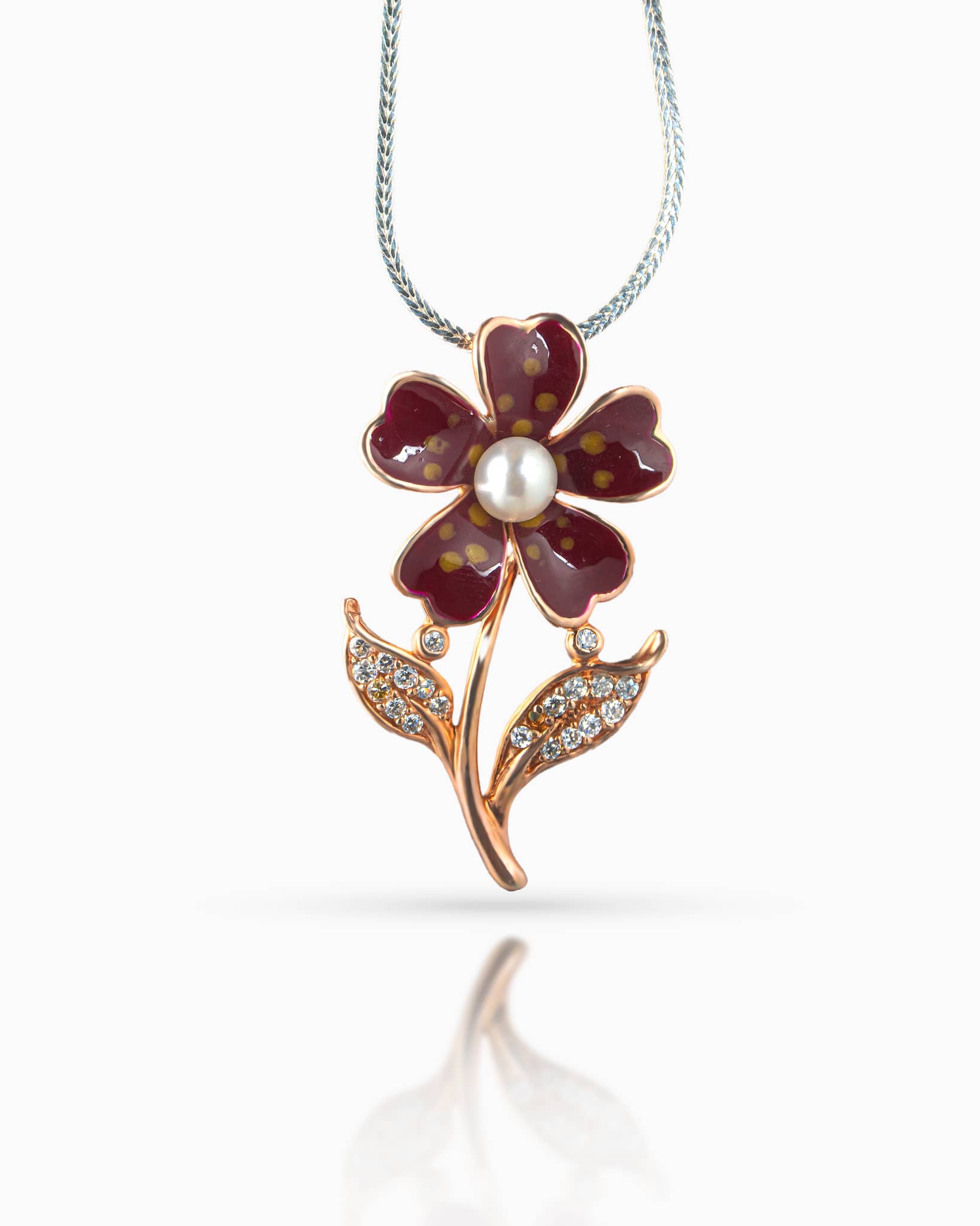 Close up of an elegant rose gold plated floral pendant showing playful maroon and yellow polka dot enamel work, a white pearl, and cz paved leaf accents resting on a white quilted surface.