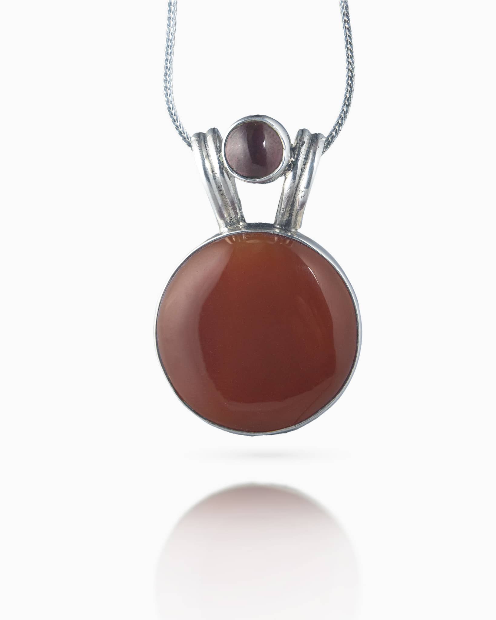 Close up of a minimalist silver pendant showing a large reddish-brown cabochon center stone and a smaller dark red stone set in a double-loop bail.