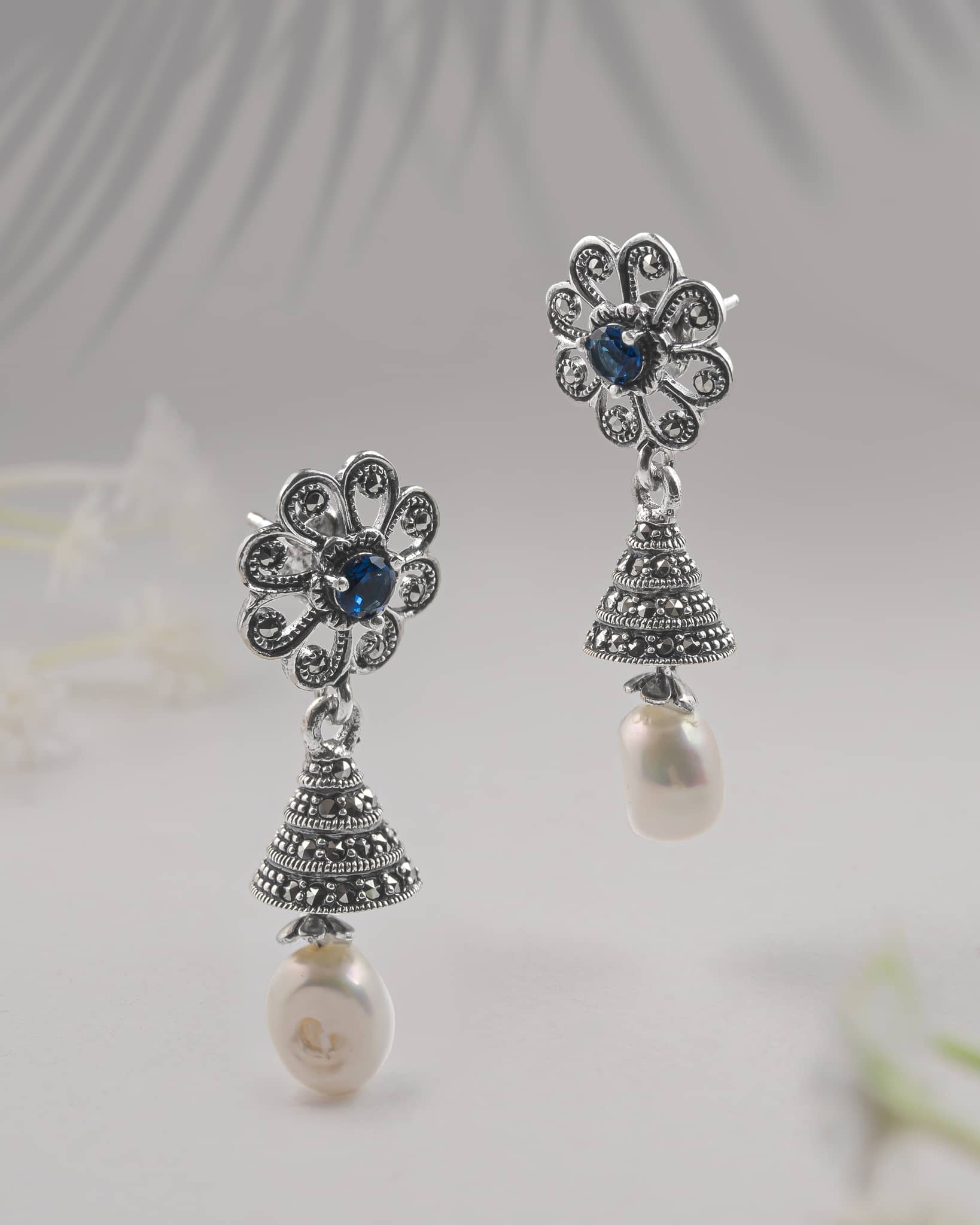 Front and side profile view of silver oxidized floral jhumkis showing the tiered bell, blue center stone, and push-back closure.