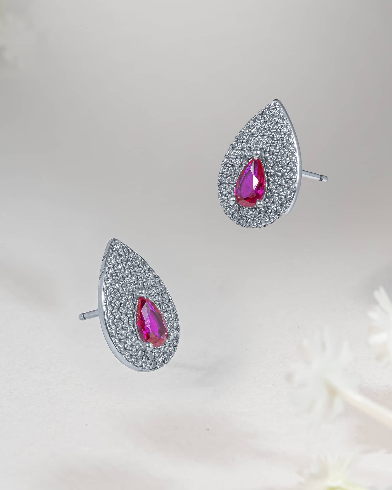 Floating angled shot of 92.5 sterling silver teardrop stud earrings featuring a faceted ruby pink cz center surrounded by a dense clear cz micro-pave halo.